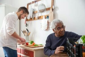 A young man provides care to an older man who is sitting down in the kitchen. They are an example of live in care that give back to charities