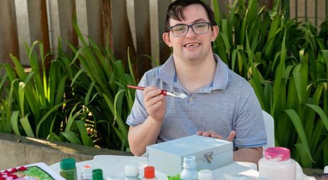 Live in care for a young man with Down’s syndrome painting a wooden box at a table
