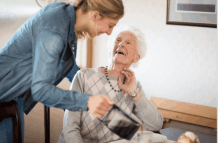 Smiling caregiver helping an older person sit comfortably on a chair while they chat together in a bright room.