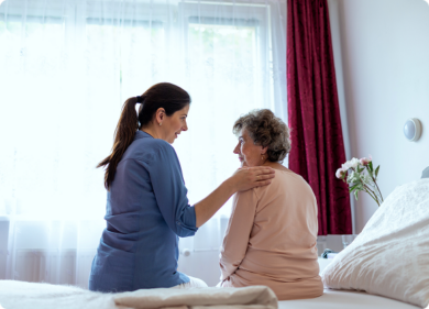 A woman assists an older woman sitting on a bed, providing support and care in a nurturing environment.