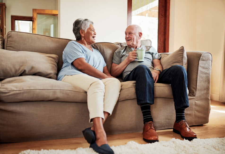 An older couple sits together on a couch in a cozy living room, smiling and enjoying each other's company