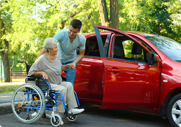 A man assists an elderly woman in a wheelchair beside a parked red car.