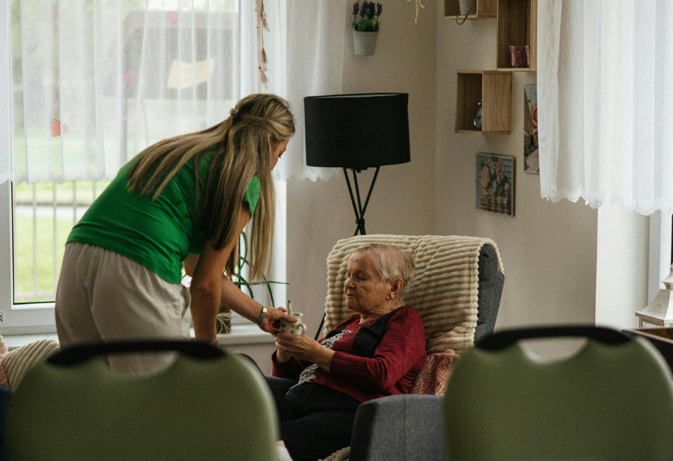 A woman assists an elderly woman seated in a chair, providing support and care in a warm environment