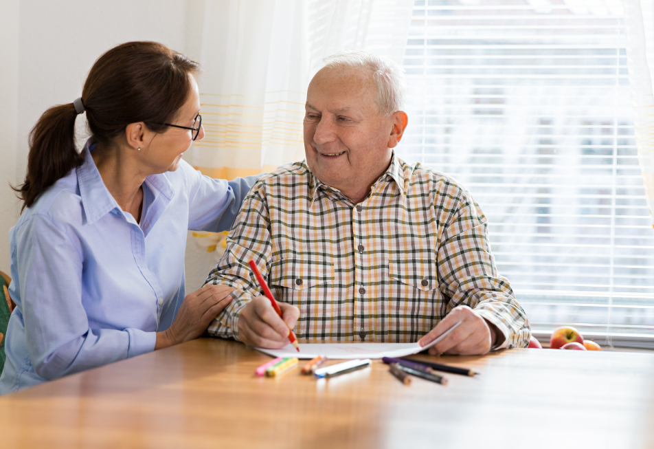 A woman assists an elderly man with his paperwork at a table, providing guidance and support